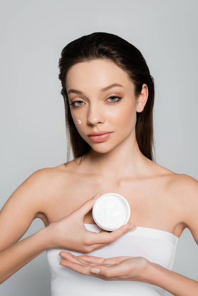 brunette young woman with bare shoulders and cream on cheeks holding container isolated on grey - Fotografie, Obrázek
