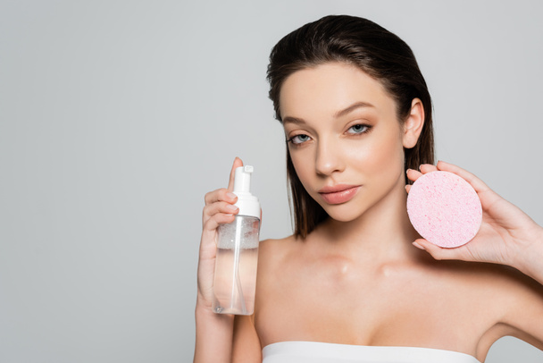 young woman holding bottle with cosmetic product near exfoliating sponge isolated on grey - Foto, immagini