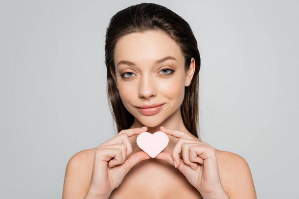 smiling young woman holding heart-shaped sponge near face isolated on grey - Φωτογραφία, εικόνα