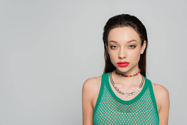 young and stylish model with red lips and necklaces posing isolated on grey - Foto, Imagen
