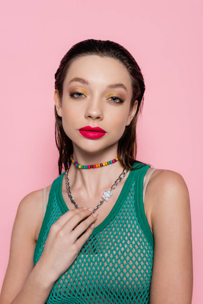 young brunette woman with bright makeup touching chain necklace while looking at camera isolated on pink  - Fotografie, Obrázek