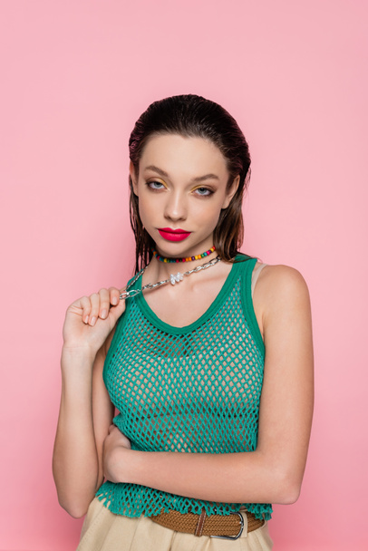 young brunette woman with bright makeup pulling silver necklace while looking at camera isolated on pink  - Foto, Imagen