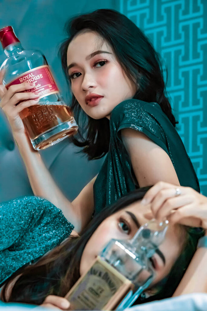 Asian women holding an alcohol bottle in their hand in the bedroom while wearing blue dresses - Photo, Image