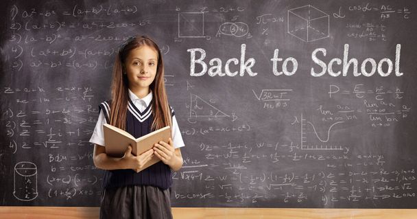 Schoolgirl with a book standing in front of a blackboard with message back to school - 写真・画像