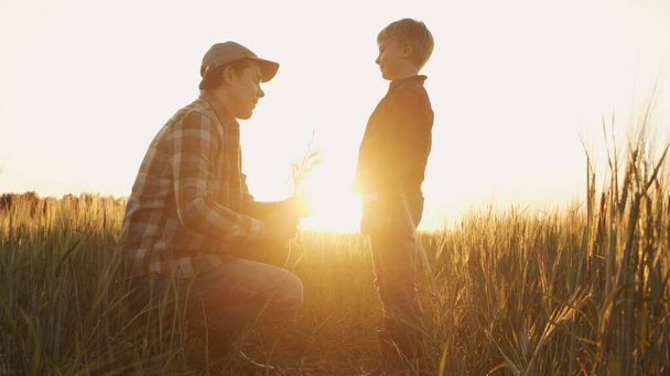 Farmer and his son in front of a sunset agricultural landscape. Man and a boy in a countryside field. The concept of fatherhood, country life, farming and country lifestyle. - Фото, изображение
