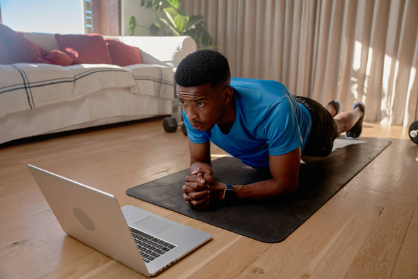Nuori afroamerikkalainen musta mies urheilija katselee harjoitusvideoita kannettavalla tietokoneellaan tekemässä online-luokkaa pitäen samalla lankku-asentoa. Kuntoilua, kuntoilua kotona. Laadukas kuva - Valokuva, kuva