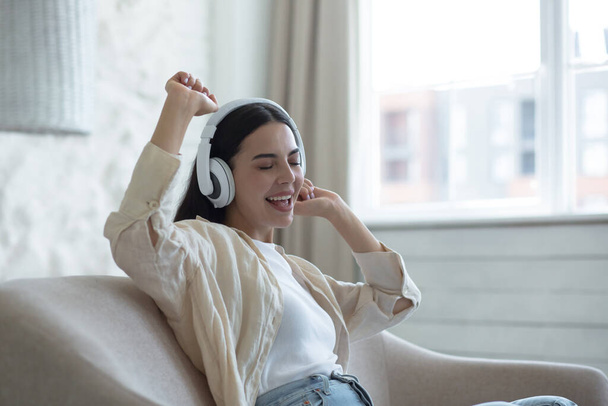 Beautiful young woman at home relaxing listening to music from white headphones sitting on the sofa in the daytime, singing along and smiling joyfully - Φωτογραφία, εικόνα