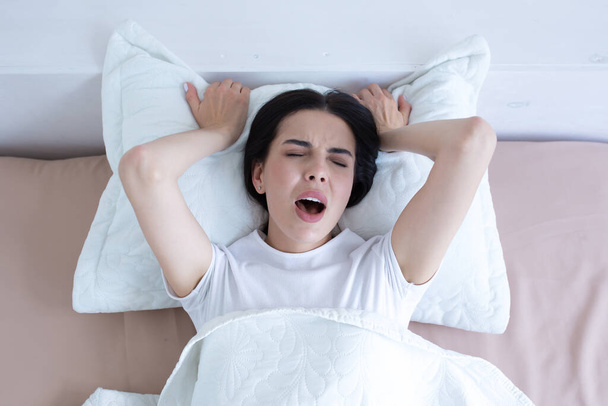 Frustrated and upset woman lying in bed alone at home crying and screaming, top view, brunette in white t-shirt pajamas - Photo, image
