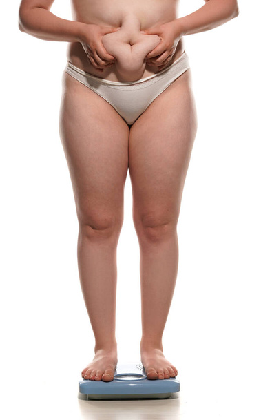 dikke buik vrouw staan op een schaal en knijpen een dikke rol op haar buik op witte achtergrond. - Foto, afbeelding