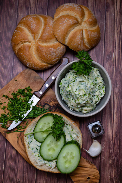 Spicy herb butter with cucumber slices on fresh bun and chopped herb ingredients on cutting board, top view - Фото, изображение