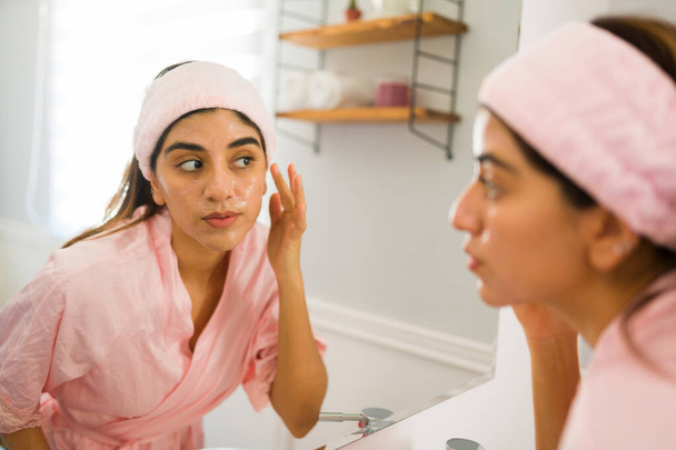 Hispanic woman in a bathrobe doing using skin care products and putting moisturizer in her face while looking in the bathroom mirror - Photo, image
