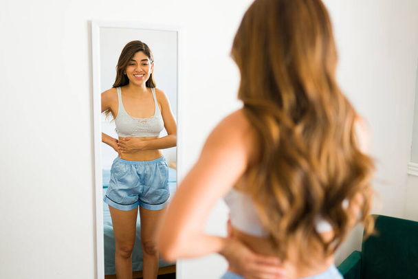 Mooie jonge vrouw met een goed gevoel van eigenwaarde te kijken naar haar slanke lichaam in de spiegel en het gevoel erg gelukkig - Foto, afbeelding