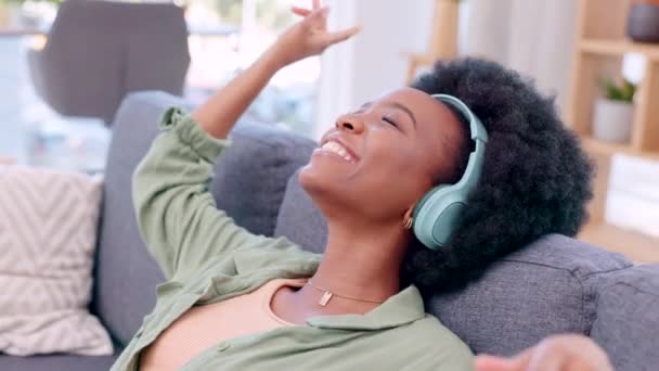 Happy, carefree and relaxed woman listening to music on headphones while sitting on a sofa in her living room at home. Young female dancing and smiling while streaming playlist songs on the weekend. - Felvétel, videó