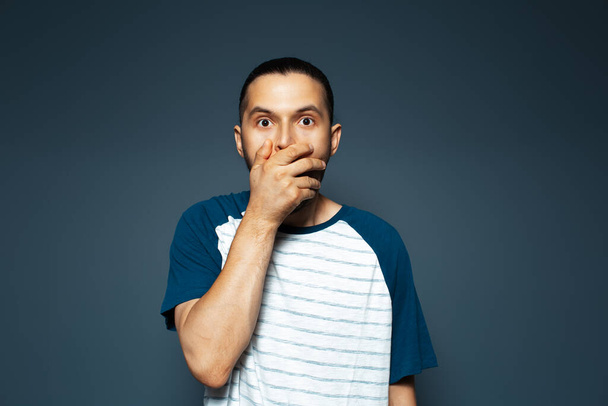 Studio portrait of guy covering mouth with hand on blue background. Wearing t-shirt. - Foto, imagen