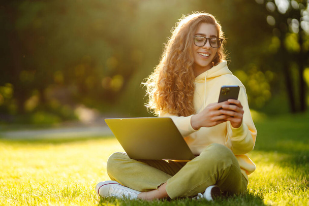 Young woman with laptop sits on the grass in the park on a sunny day. Online training, remote work and communication in social networks. Business, blogging, freelance. - Valokuva, kuva