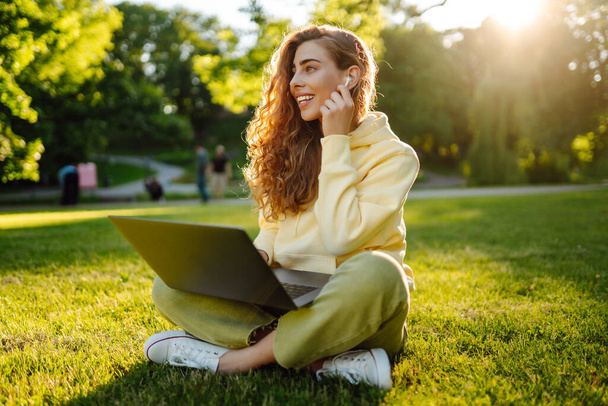 Young woman with laptop sits on the grass in the park on a sunny day. Online training, remote work and communication in social networks. Business, blogging, freelance. - Foto, Imagem