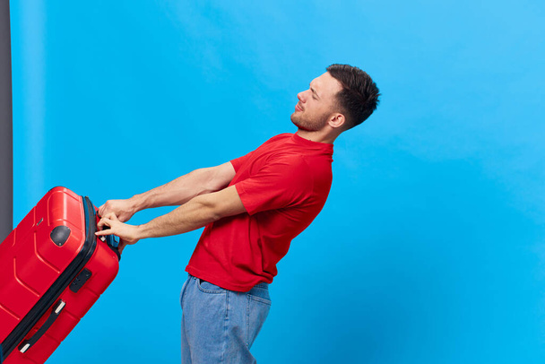 Втомлений незадоволений засмаглий красивий чоловік у червоній футболці, який готується до ділової поїздки, тримає важку валізу, позуючи ізольовано на синьому студійному фоні. Копіювати пробіл Банерний макет. концепція подорожей - Фото, зображення