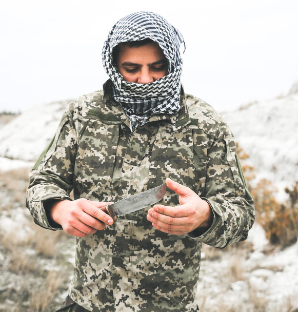 Soldier is standing in the camouflage uniform and checkered keffiyeh shemagh bandana. Man with knife is outdoors in the abandoned deserted place. - Foto, Imagen