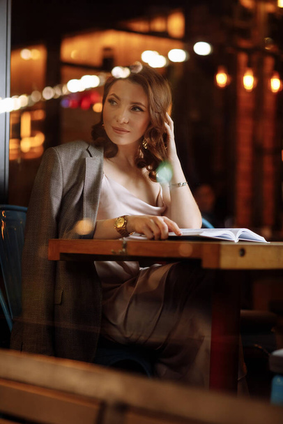 portrait of young beautiful woman is reading paper book in cafe in dusk. girl is online study, remote work - Foto, Imagem