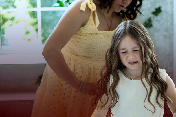 stepmother combing her daughters hair. not own mother who hurts. A girl in festive dress with a displeased expression on her face. Tangled baby hair. The concept of pain from combing curls daughter - Фото, зображення