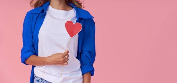 A young woman in a blue shirt holds a red paper heart in front of her chest. Pink studio background. The concept of women's health and diseases of the cardiovascular system - 写真・画像