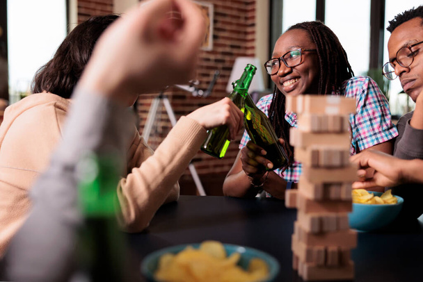 Monikulttuuriset onnelliset ystävät nauttivat alkoholijuomien juomisesta yhdessä pelatessaan yhteiskunnan pelejä. Monietninen iloinen ihmiset istuvat kotona olohuoneessa rentoutuen vapaa-ajan toimintaa. - Valokuva, kuva