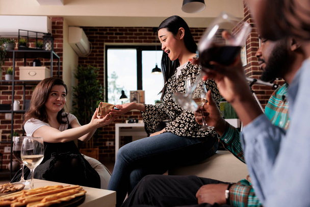 Diverse group of friends giving out each other gifts while having wine party at home. Young adults enjoying time together while drinking alcoholic beverages and exchanging presents. - 写真・画像
