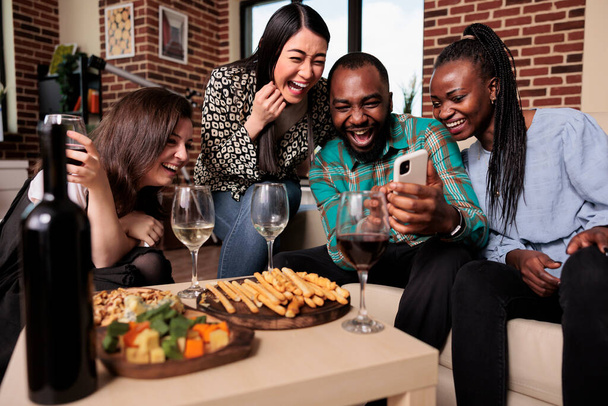 Různorodá skupina přátel na vinařské party slaví narozeniny při sledování legrační klipy na smartphonu. Šťastní mladí lidé, kteří si dávají sklenku vína a jedí svačinky, zatímco spolu tráví čas. - Fotografie, Obrázek