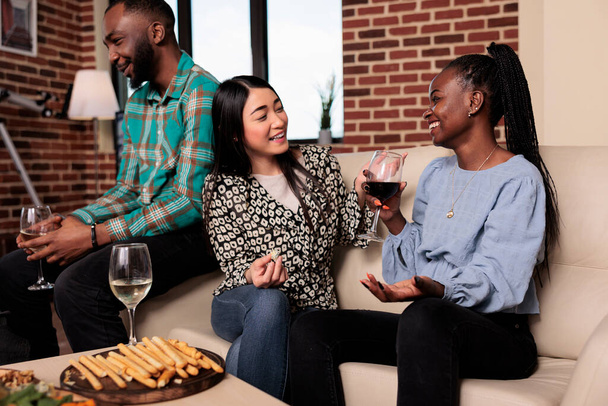 Multiethnic joyful women at wine party talking while sitting on sofa. Happy smiling diverse friends sitting in living room while enjoying alcoholic beverages together while celebrating birthday event. - Фото, изображение