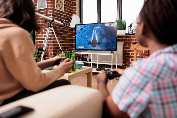 Diverse close friends in living room sitting on sofa while playing space sim videogame on gaming device. People sitting at home while enjoying fun leisure activity with entertainment console device. - Zdjęcie, obraz