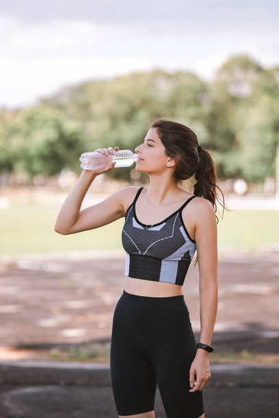 Beautiful Caucasian girl athlete in dressed in sportswear: shorts and top, sneakers with fitness tracker drinks water on stadium in summer. Happy female outdoor sport and healthy lifestyle concept - Zdjęcie, obraz