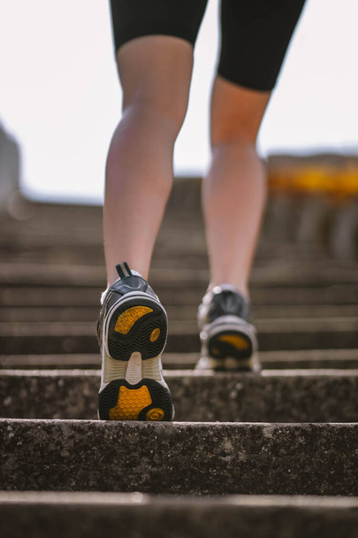 Closeup photo of athlete girl's legs dressed in sportswear with sneakers going up stairs outside in city abandoned stadium . Happy young female sport, travelling, ecology, healthy lifestyle concept - Foto, Imagen