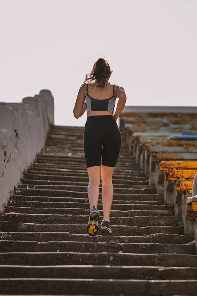Beautiful Caucasian girl athlete dressed in sportswear: shorts and top, sneakers, fitness tracker runs on stadium stairs in summer. Happy young female outdoor sport, workout, healthy lifestyle concept - Foto, immagini