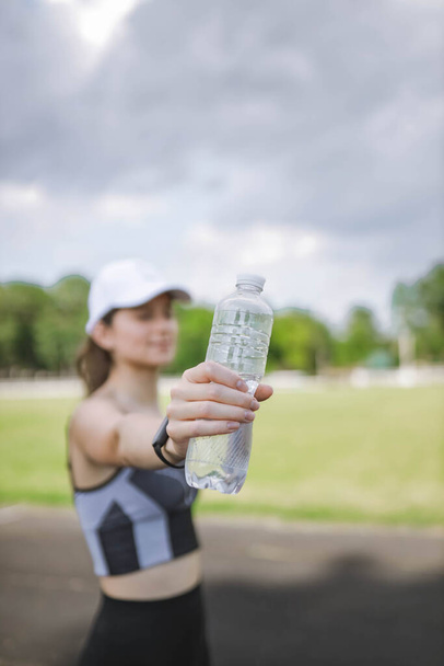 Beautiful Caucasian girl athlete in dressed in sportswear with fitness tracker on stadium does fitness and holds plastic water bottle. Selective focus. Happy female sport and healthy lifestyle concept - Valokuva, kuva