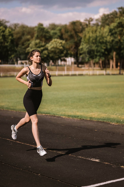 Concentrated Caucasian girl athlete dressed in sportswear: shorts and top, sneakers with fitness tracker running on track at city stadium in hot summer. Happy female outdoor workout  sport concept - Foto, afbeelding