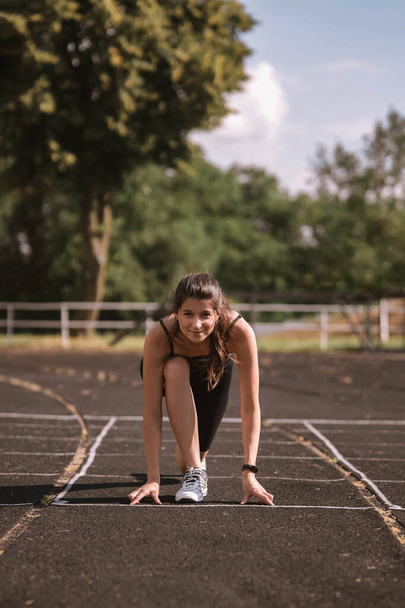 Concentrated Caucasian girl athlete in dressed in sportswear: shorts and top, sneakers with fitness tracker preparing for run on track at stadium in summer. Happy female outdoor sport concept - Foto, immagini