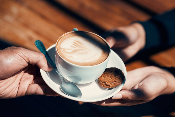 Closeup of hands holding and giving a cappuccino or flat white to a customer at a cafe. Hot beverage with creative plant image in milk foam. A morning coffee drink made by a barista in a restaurant - Photo, image