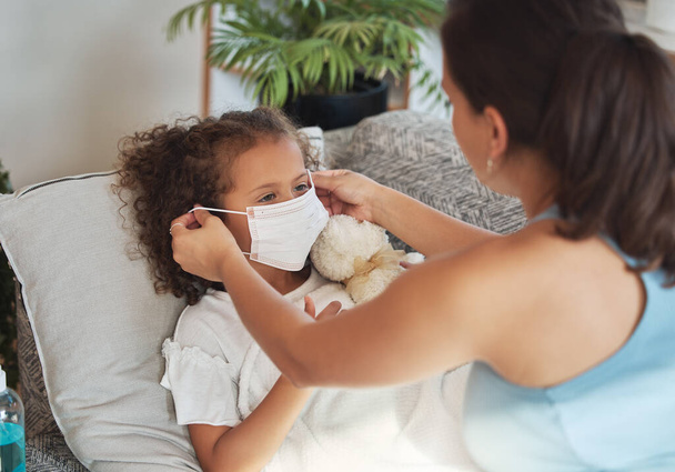 Health care for sick, tired little girl with covid wearing face mask while resting on a sofa at home with worried mother. Anxious parent helping her unwell daughter to protect and stop virus spread. - Foto, Imagen