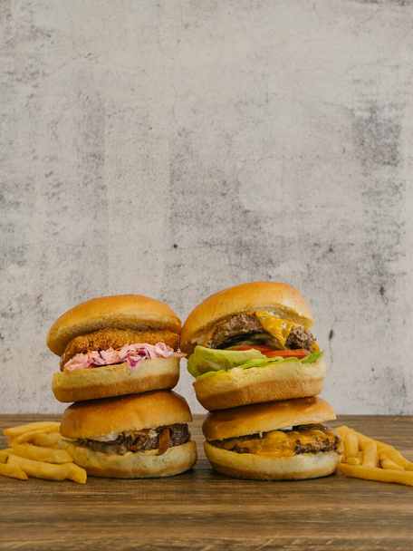 vier hamburgers opgestapeld twee met frietjes aan de zijkant op houten tafel met kopieerruimte op betonnen muur - Foto, afbeelding