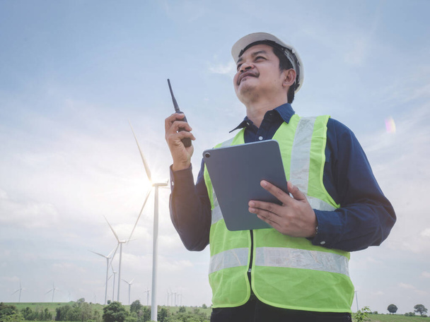 Asian engineer man stand holding tablet front the wind turbines generating electricity power station with talking by walkie -talkie . Concept of sustainability development by alternative energy - Φωτογραφία, εικόνα