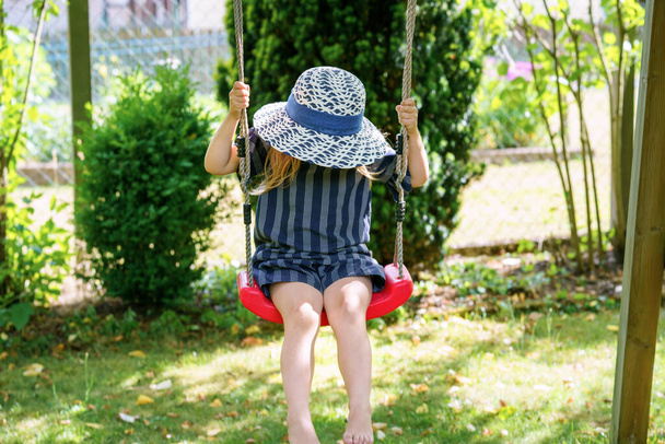 Happy little preschool girl having fun on swing in domestic garden. Smiling positive healthy child swinging on sunny day. Preschool girl laughing and crying. Active leisure and activity outdoors - Foto, Imagem