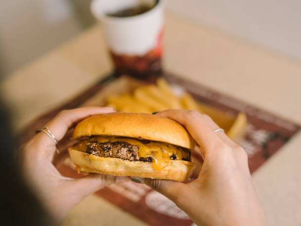 Lady POV holding single cheese burger with tray of fries and soda in background - Foto, imagen