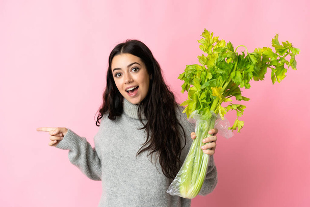Young caucasian woman holding a celery isolated on blue background surprised and pointing finger to the side - Foto, Imagen