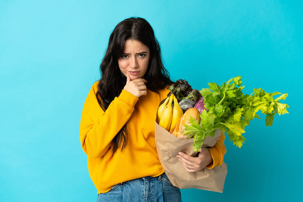Mujer joven sosteniendo una bolsa de la compra de comestibles aislado en el pensamiento de fondo azul - Foto, Imagen