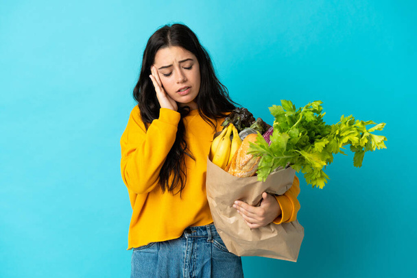 Young woman holding a grocery shopping bag isolated on blue background with headache - Photo, Image