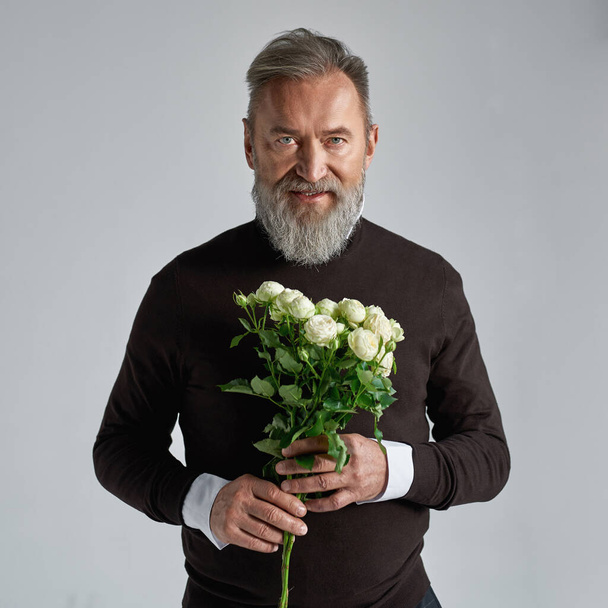 Front view of grey hair caucasian man with bouquet of white roses. Fashionable bearded male. Present for romantic meeting, Vaentines Day and 8 march. Grey background. Studio shoot. Copy space - Foto, Imagen
