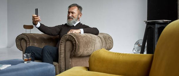 Senior smiling elegant caucasian man taking selfie on smartphone in armchair in spacious apartment. Stylish bearded pensioner. Modern senior male lifestyle. Domestic entertainment and leisure - Fotó, kép