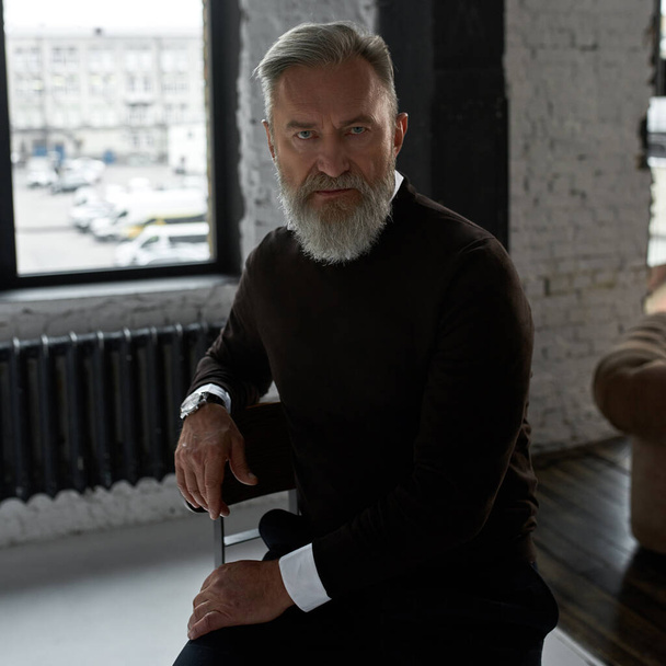 Grey hair confident caucasian man sitting on high chair and looking at camera in spacious flat. Fashionable bearded pensioner wearing casual clothes. Concept of modern elderly male lifestyle. Daytime - Fotografie, Obrázek