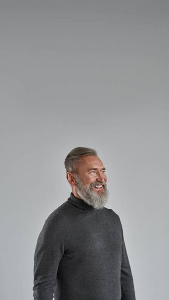 Elderly smiling caucasian man looking away. Trendy bearded pensioner wearing sweater. Concept of modern senior male lifestyle. Isolated on grey background. Studio shoot. Copy space - Photo, image