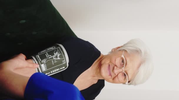 Valkoihoinen sairaanhoitaja, yllään tummansininen univormu ja kasvonaamio, istuu vanhemman naispotilaan kanssa sohvalla ja mittaa verenpainetta. Pystysuora. Laadukas 4k kuvamateriaalia - Materiaali, video